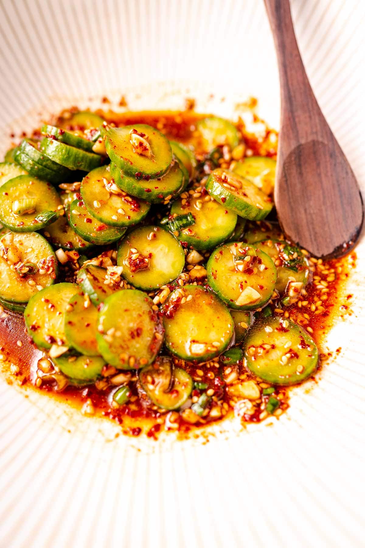 A wooden spoon stick out of an Asian cucumber salad resting in a large white bowl.