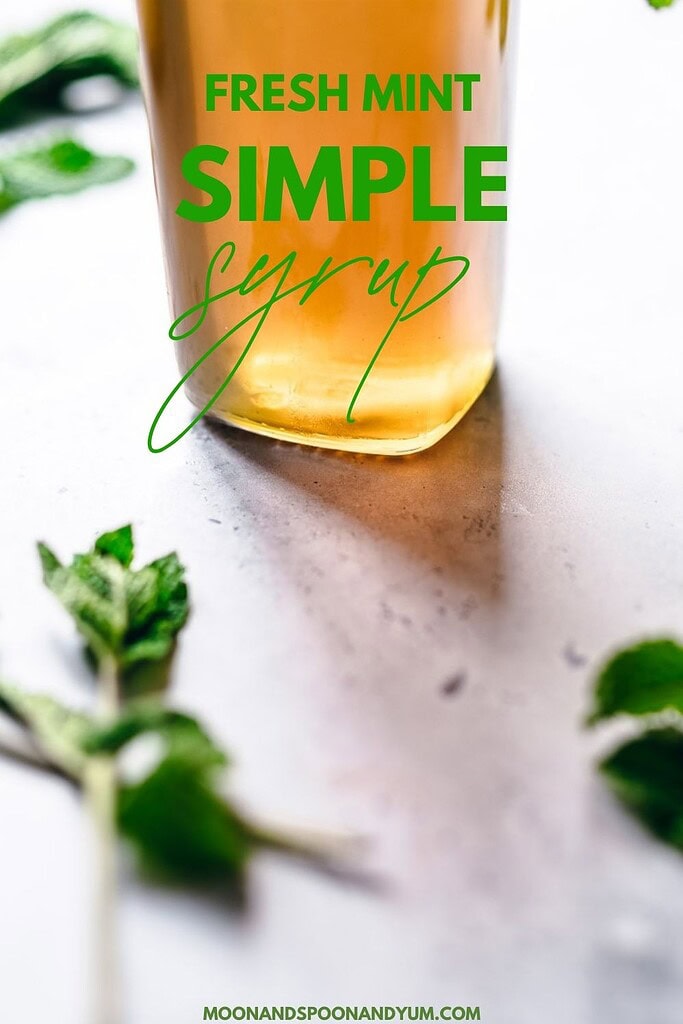 A jar of mint simple syrup sits on a light surface, surrounded by scattered mint leaves; text reads “Fresh Mint Simple Syrup.”.