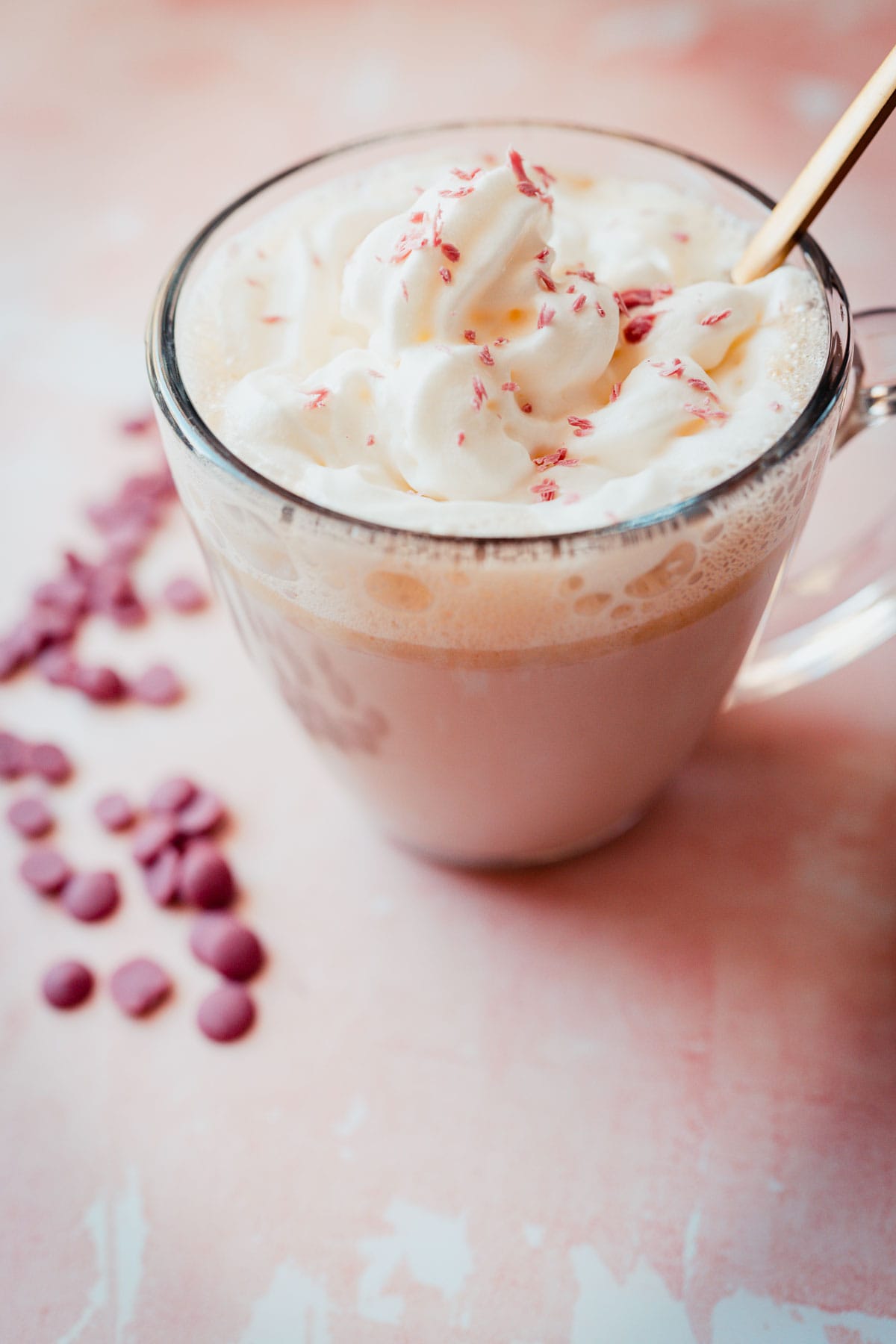 Side view of a clear glass mug filled with a pink liquid and topped with whipped cream and pink chocolate shavings.