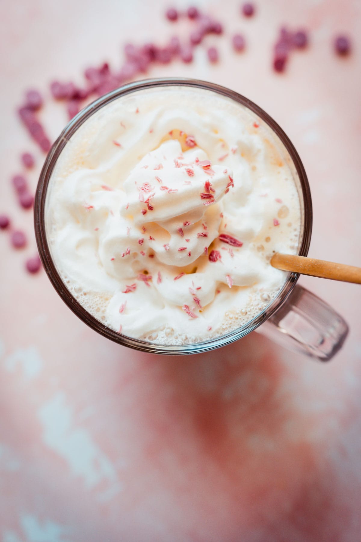 A gold spoon juts out of a clear glass mug of hot chocolate topped with whipped cream and pink chocolate shavings.