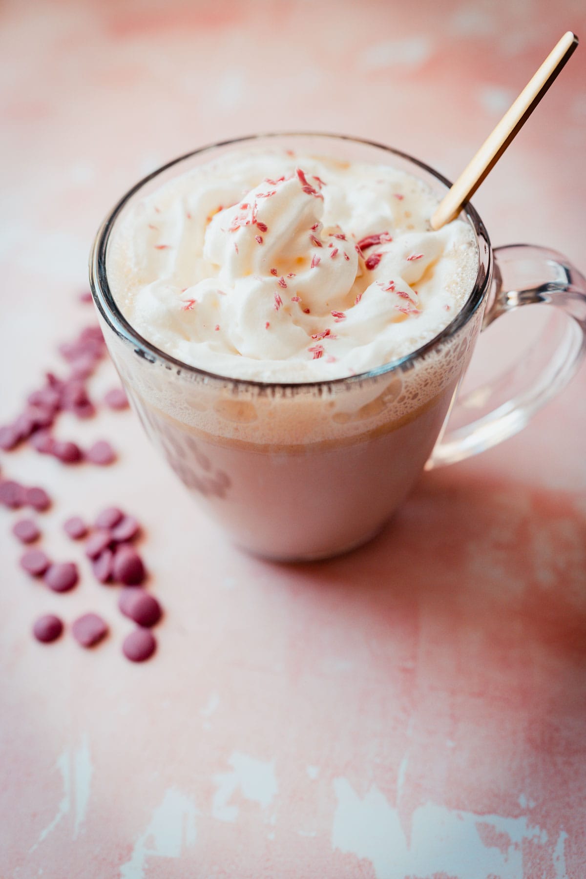 Side view of a clear glass mug of pink hot chocolate topped with white whipped cream and small pink colored chocolate shavings.