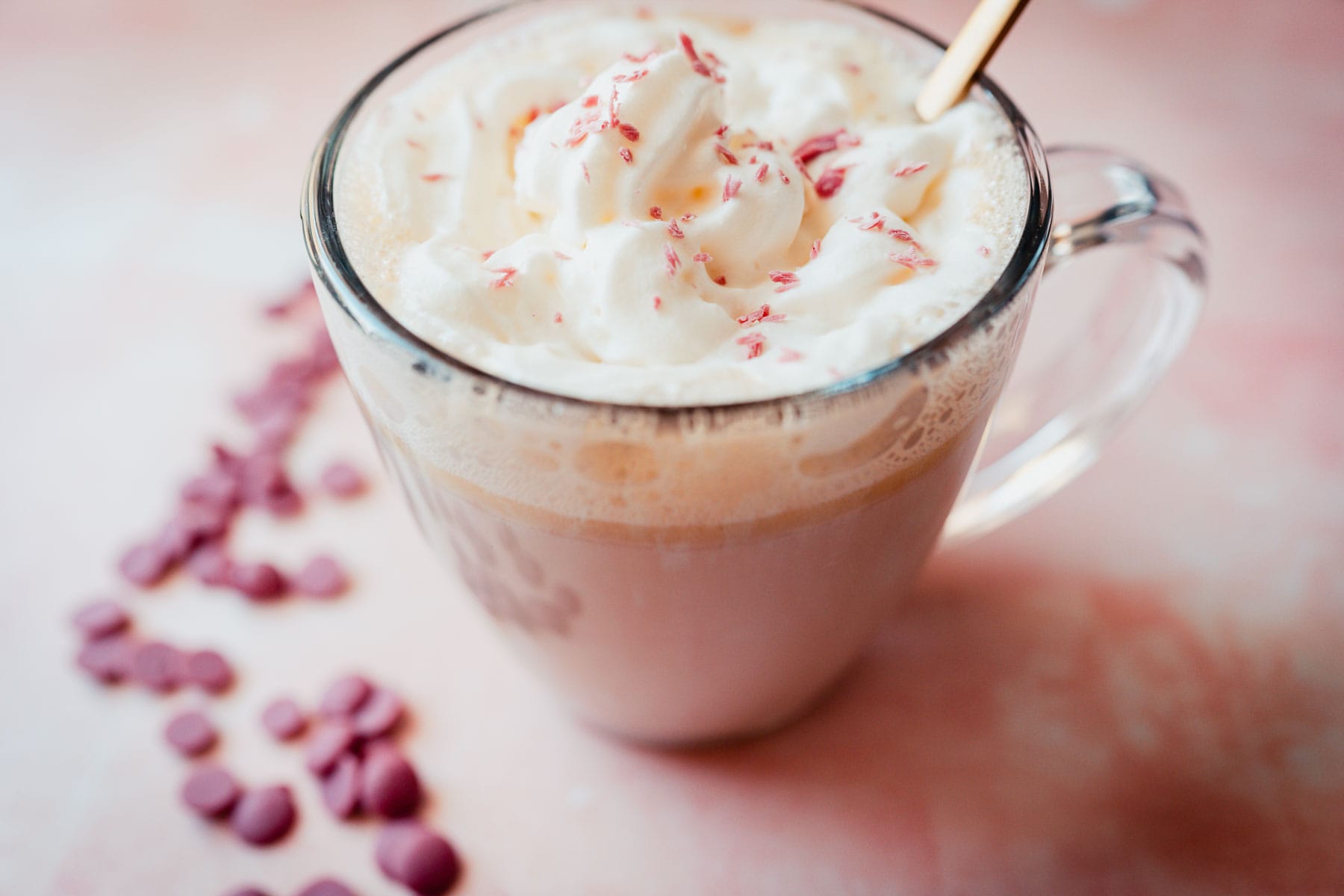 Side view of large glass mug of pink colored hot cocoa topped with frothy white whipped cream and shaved pink chocolate.