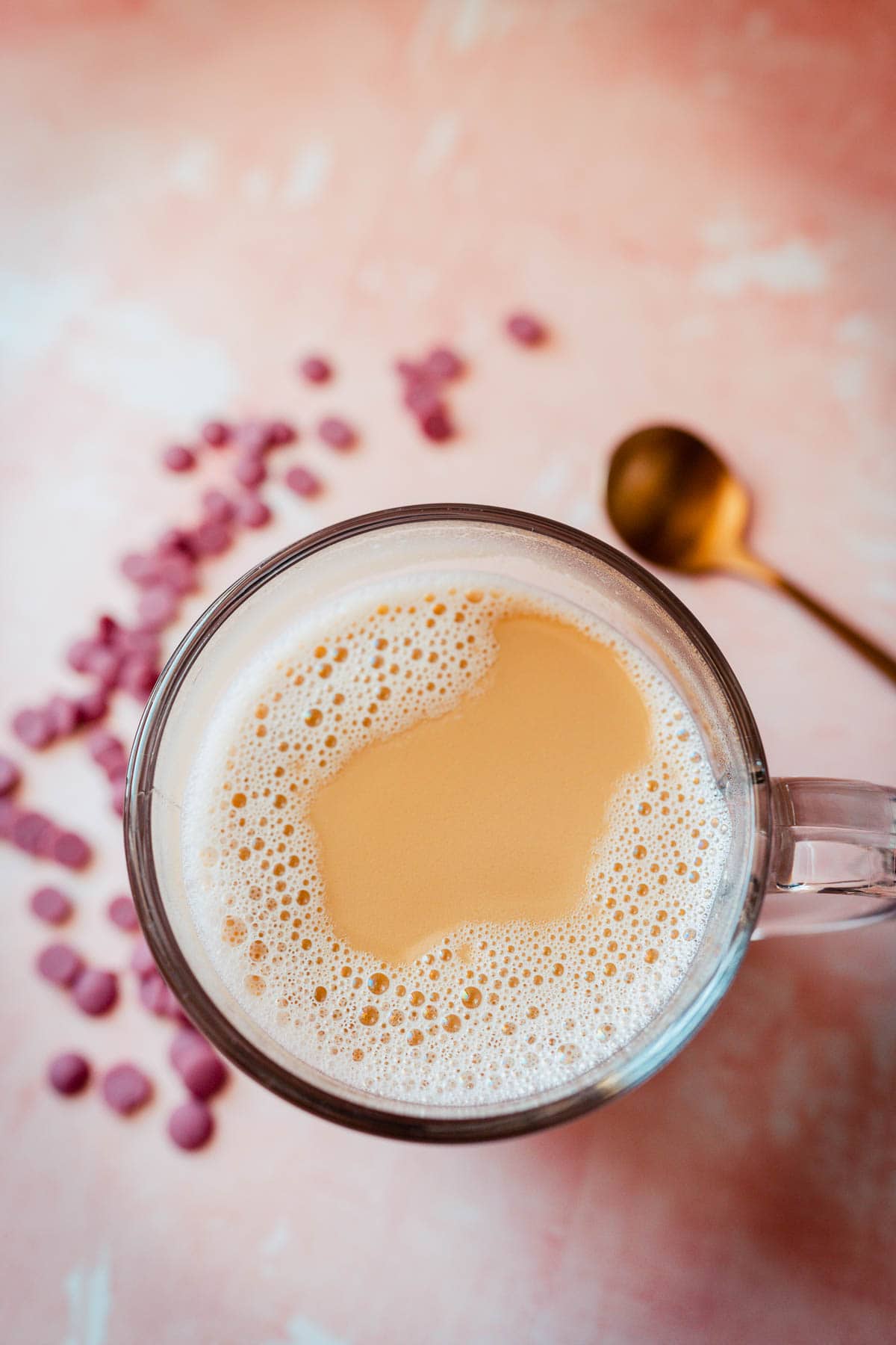 Top view of a clear glass mug of homemade ruby hot chocolate resting on a pink table next to a gold spoon.