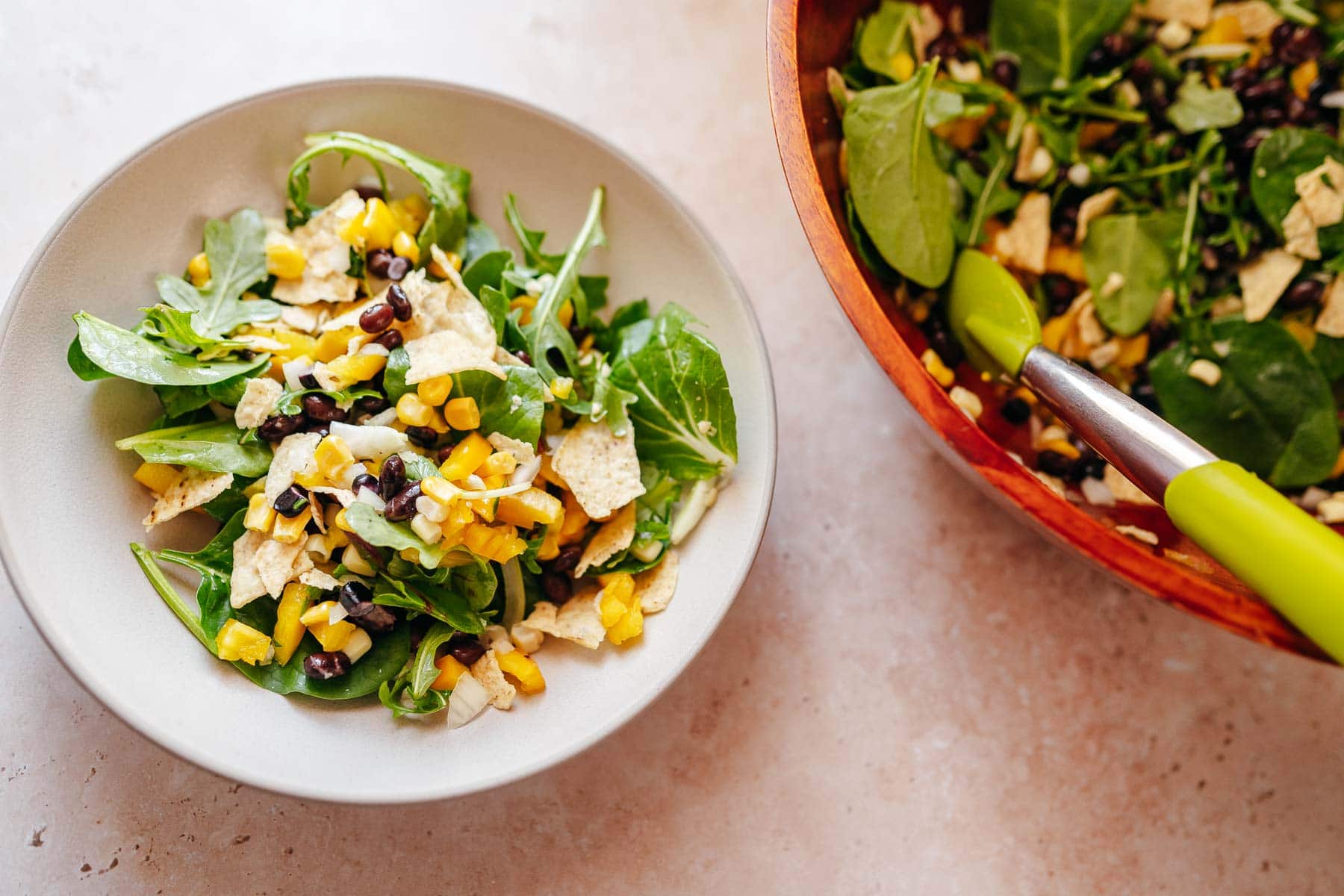 A white ceramic bowl of salad resting next to a large wood salad bowl with green tongs.