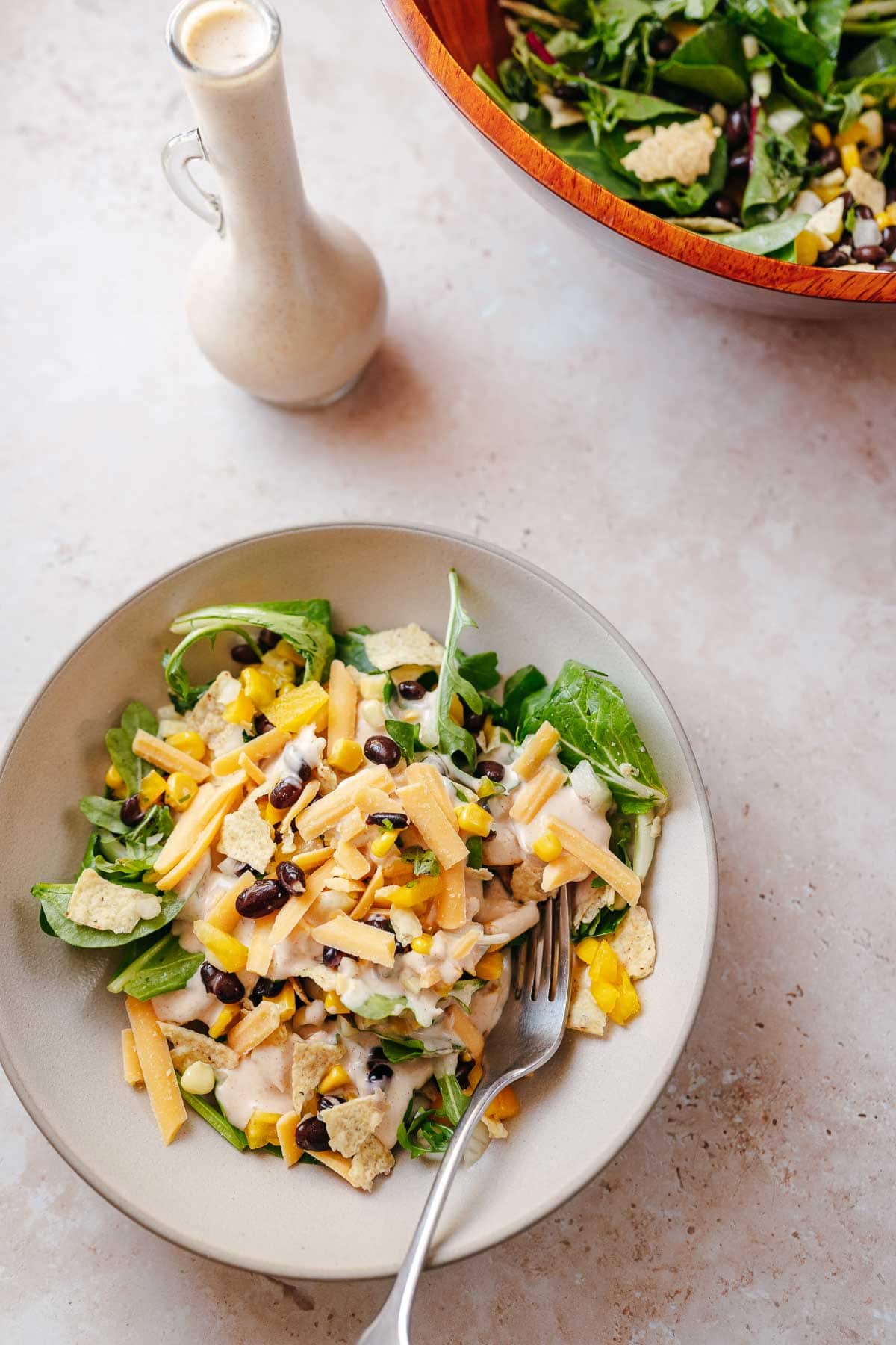 A white ceramic bowl filled with homemade southwest salad resting next to a clear glass bottle of creamy dressing and large wooden salad bowl.