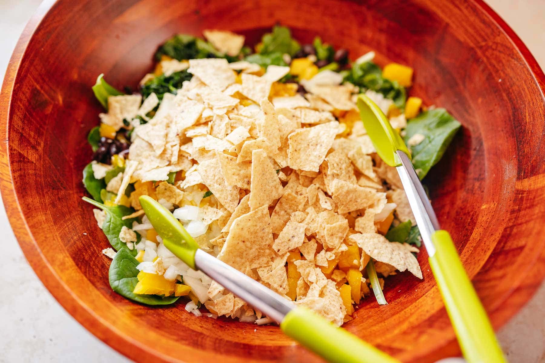 A large salad with various salad ingredients topped with tortilla strips and green tongs.