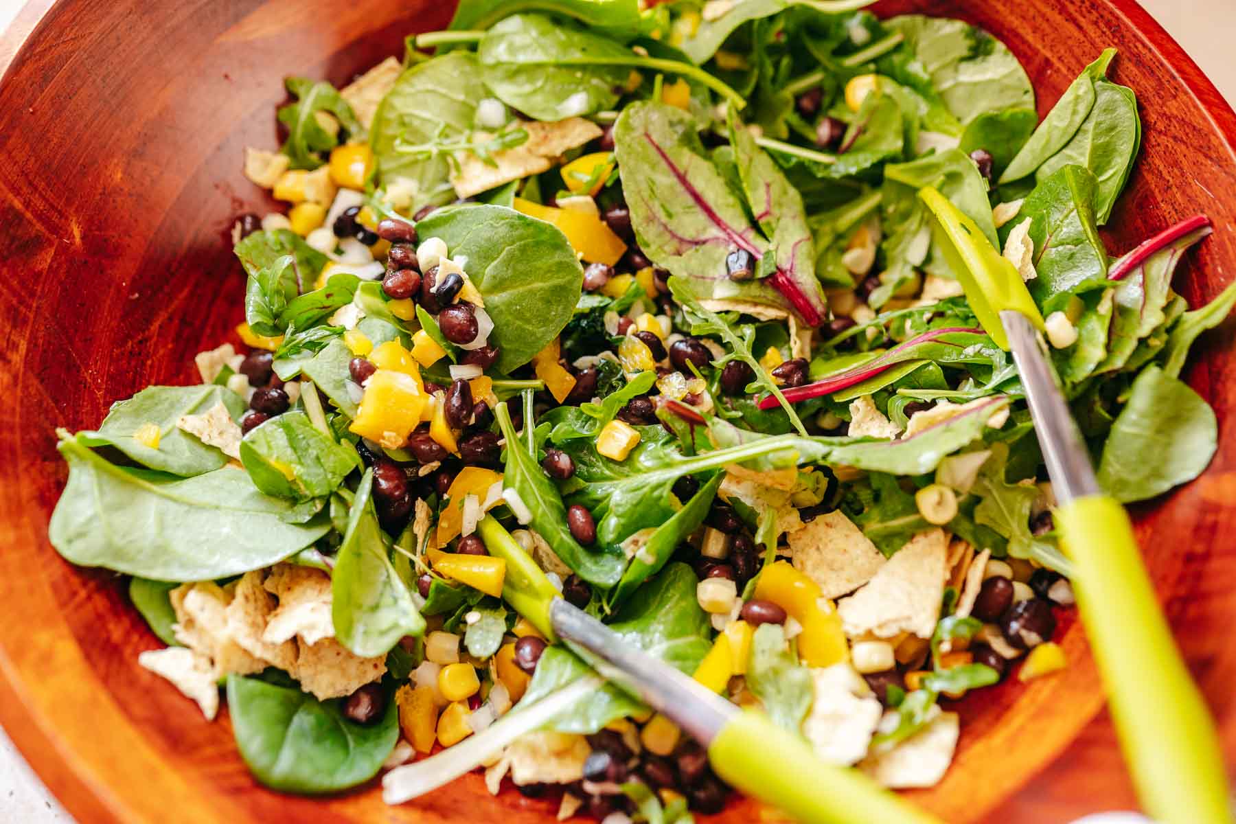 Green tongs tossing a salad in a large wooden salad bowl.