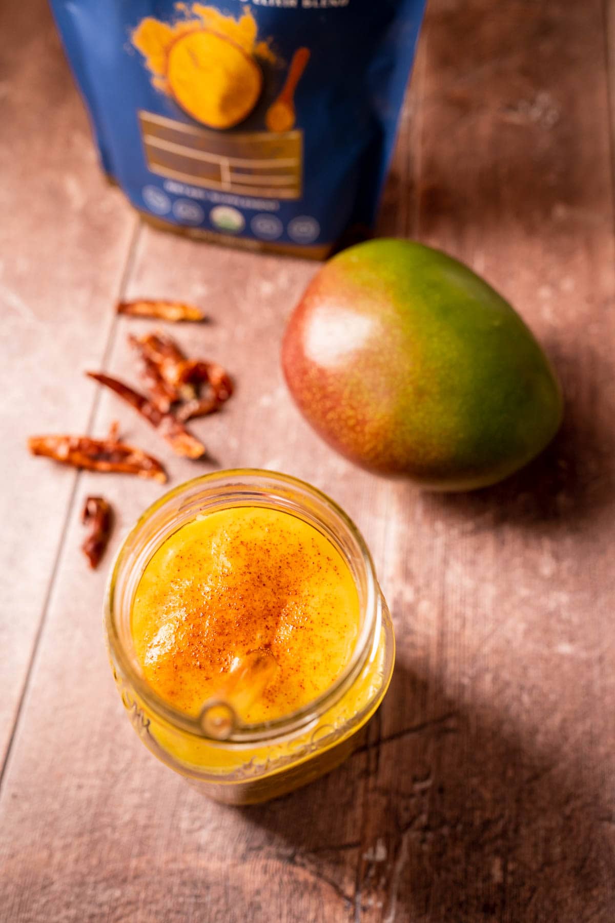 An orange smoothie resting on a wood table next to a fresh mango and dried cayenne peppers.