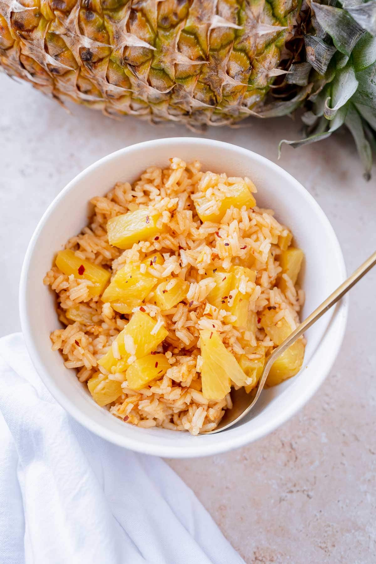 A gold spoon sticks out of a white bowl of pineapple and rice.