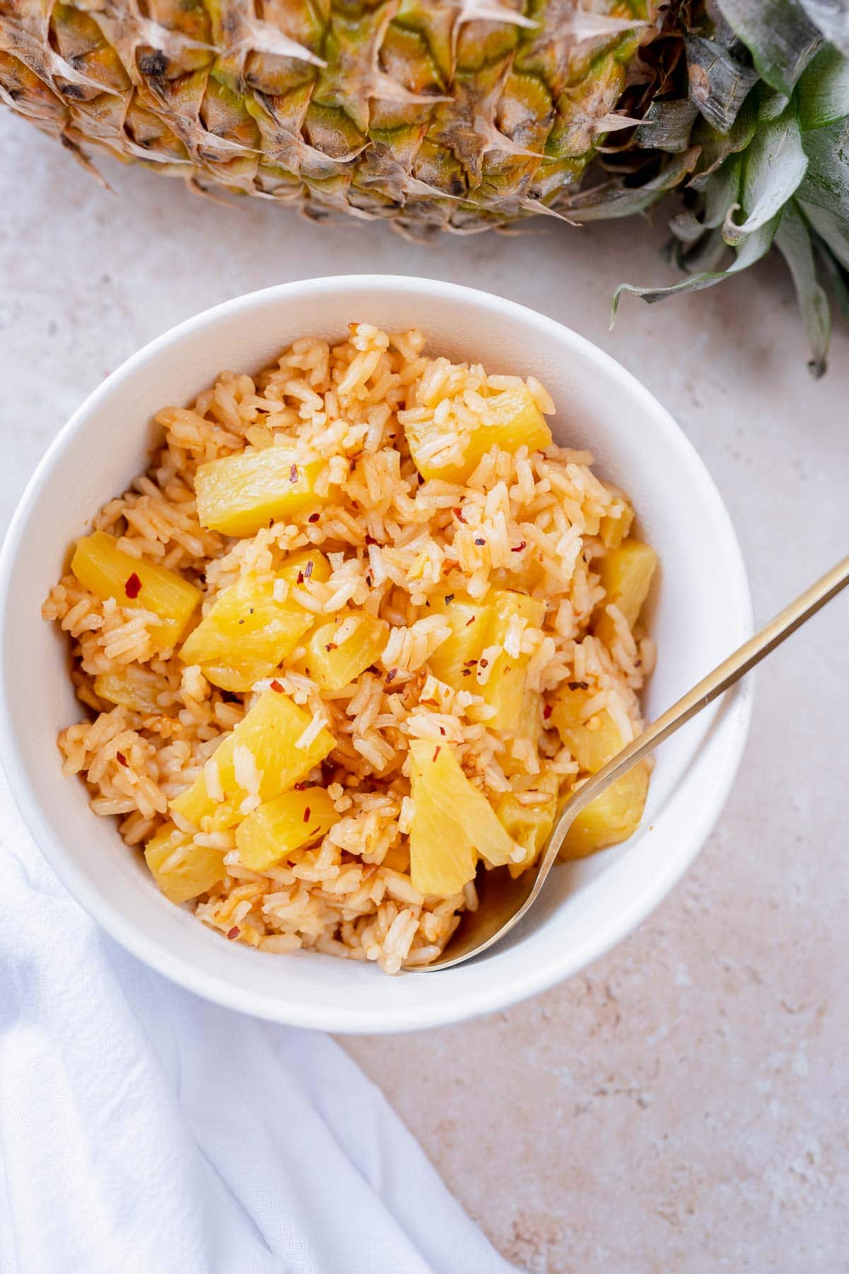 A golden spoon juts out of a white bowl filled with white rice, pineapple and red pepper flakes.