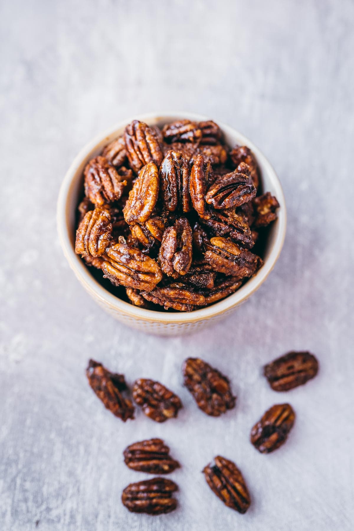 A white ceramic bowl filled with spicy pecans rests on a gray table scattered with pecans.