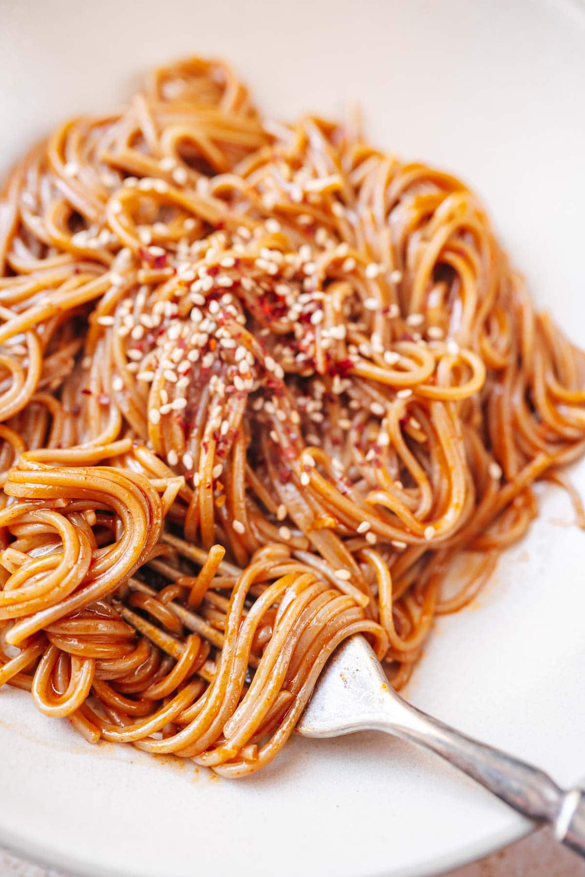 A bowl of gochujang noodles in a tan ceramic bowl with a silver fork.