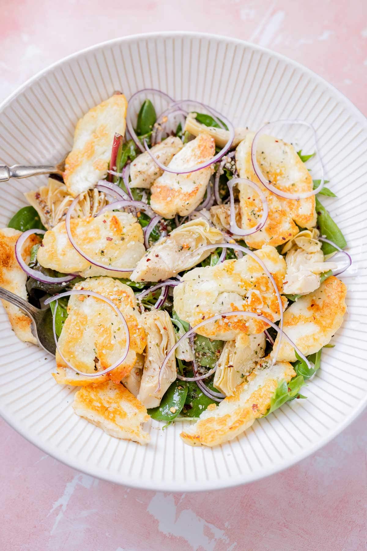 A large white bowl of salad with greens, red onion slices, seeds and crispy halloumi cheese slices.