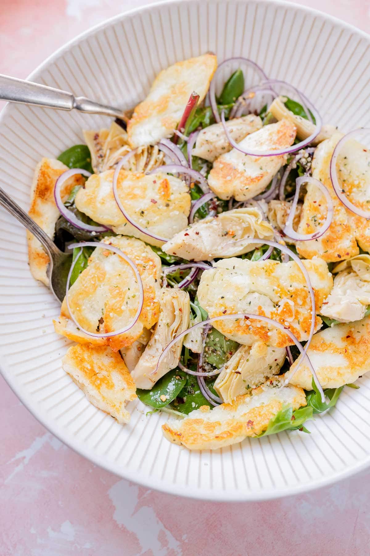 A large white bowl of halloumi, artichoke hearts, salad greens, seeds and red onion slices.