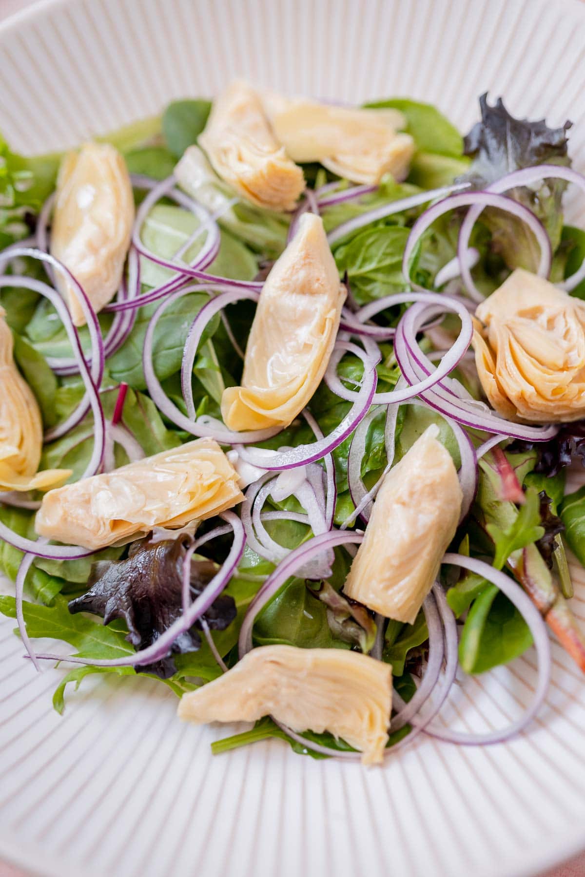 A large white bowl of salad greens, onion slices, and artichoke hearts.