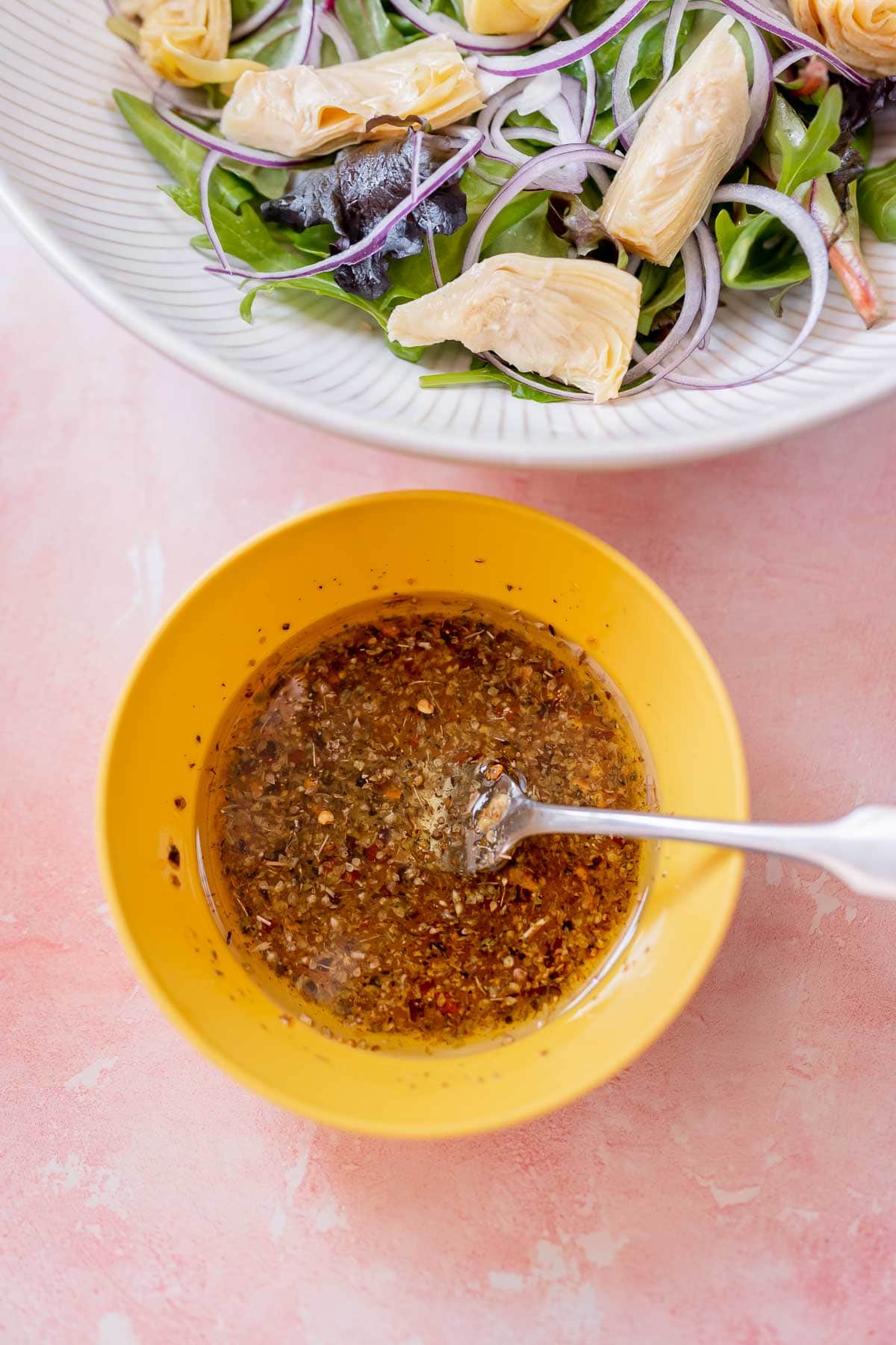 A small yellow bowl of salad dressing ingredients resting on a pink table next to a large bowl of salad.