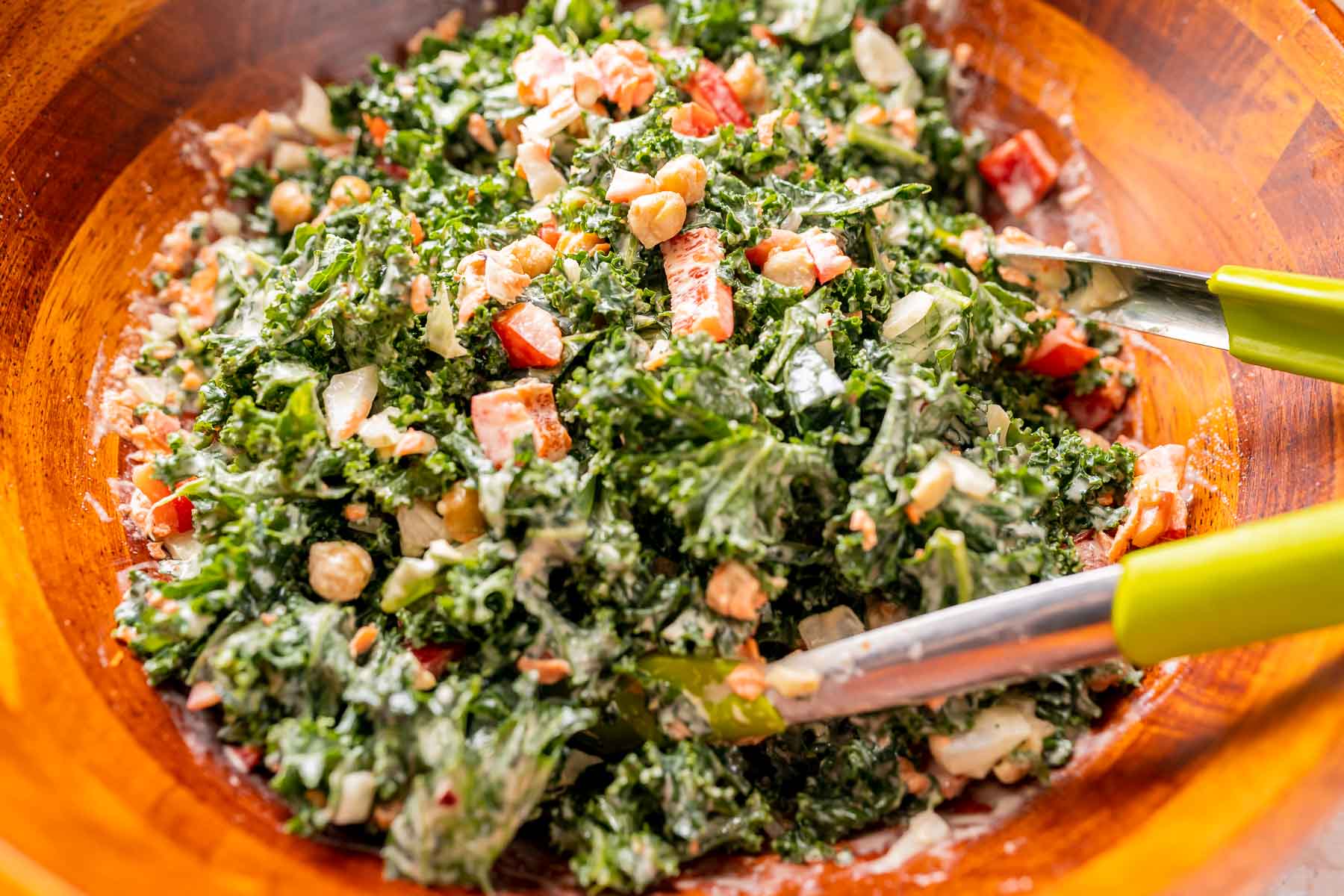 Green tongs reach into a large wood bowl of kale salad.