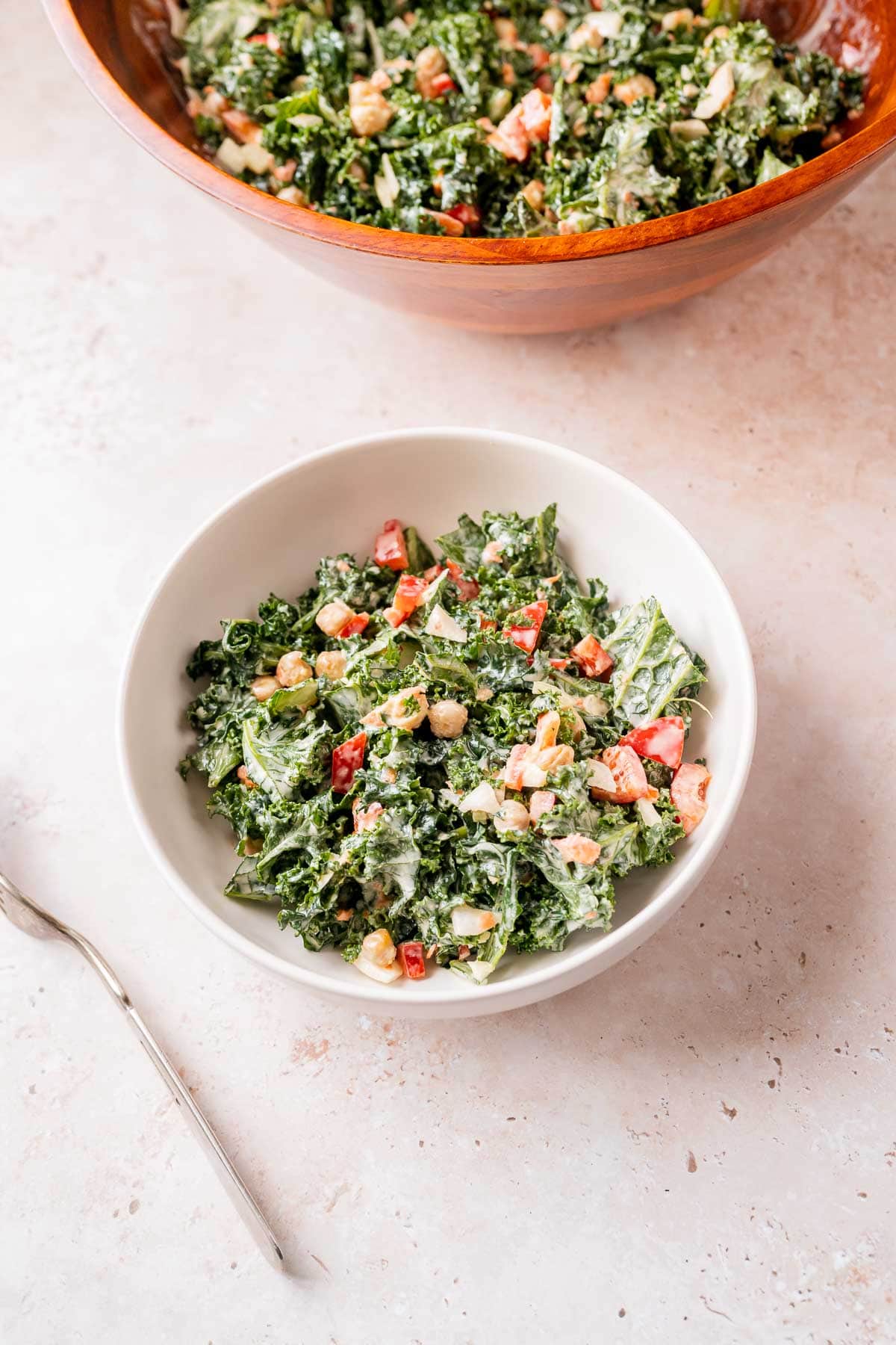 A white bowl of kale salad rests on a tan table next to a silver fork and a large wooden salad bowl.