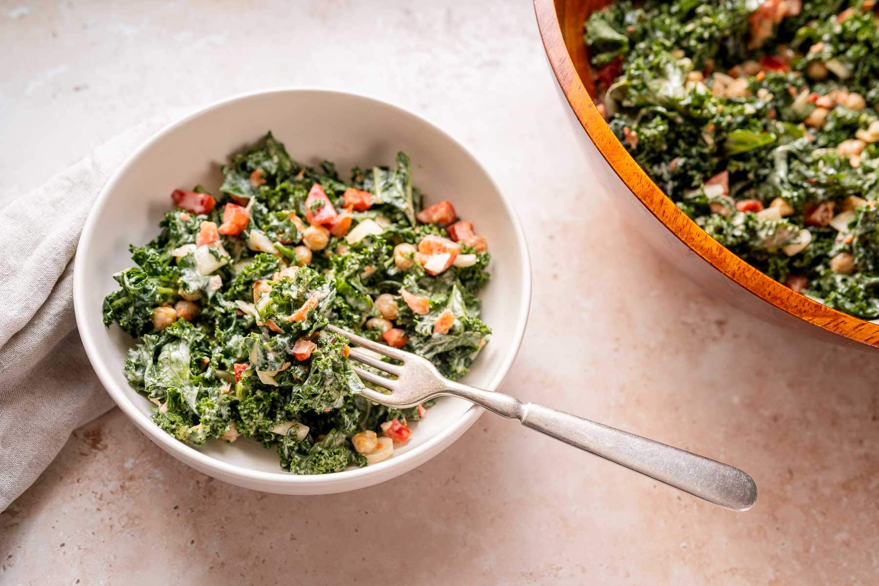 A small white bowl of kale salad rests on a tan table next to a large wood salad bowl.