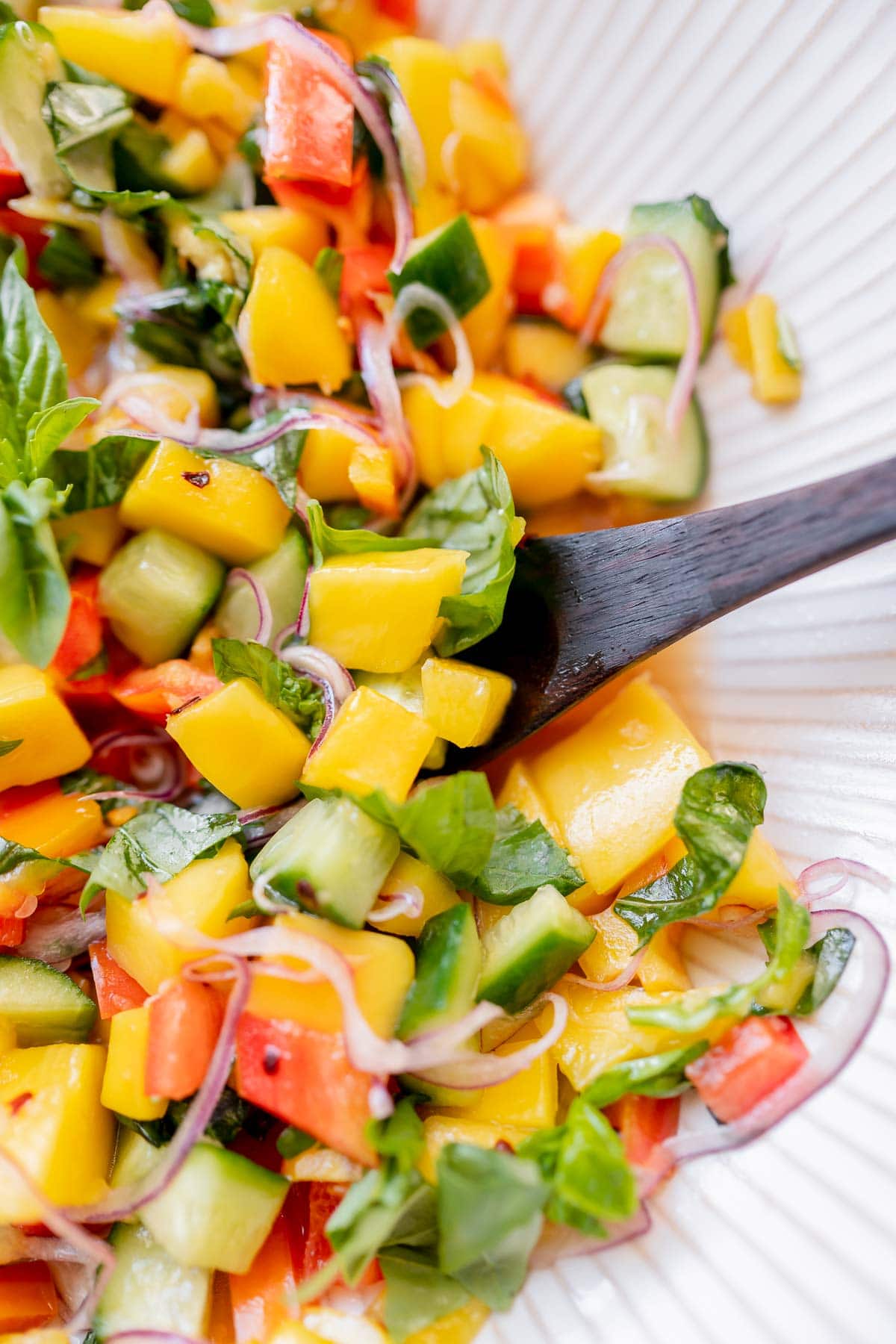 A wooden spoon scoops mango salad out of a large white bowl.