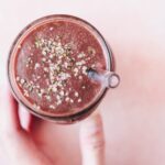 A hand holds a glass cup filled with a thick chocolate-colored acai smoothie, topped with sprinkled seeds, and a clear straw, set against a pale pink background.