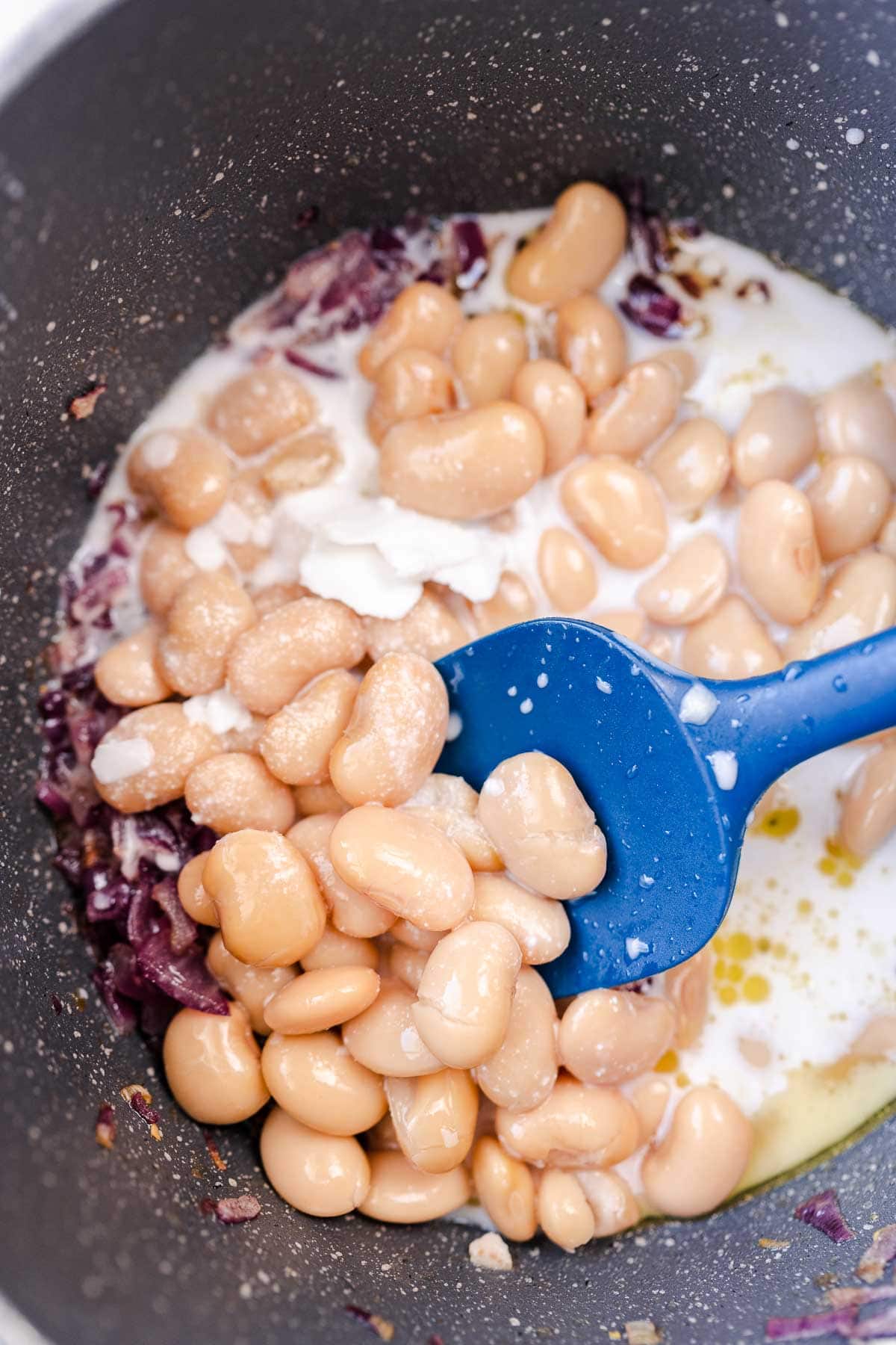 A blue spatula dips into a bot of uncooked butter beans and other ingredients.