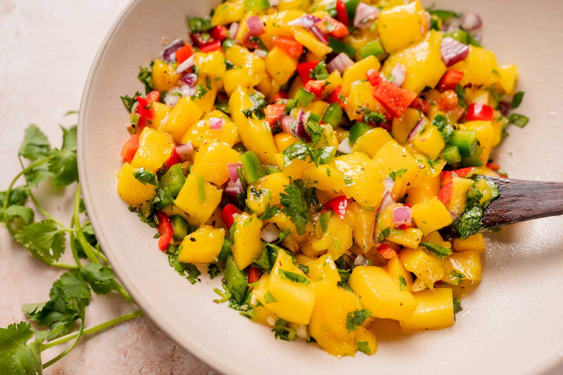 A wooden spoon rests in a bowl of bright and colorful mango salsa.