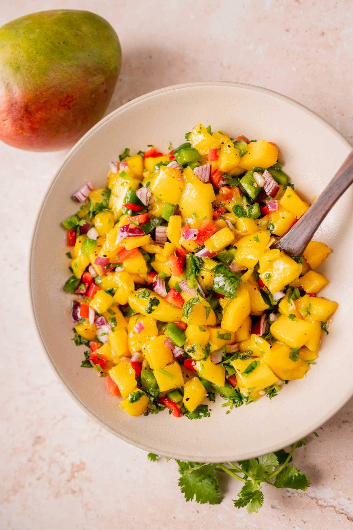 A large ceramic bowl of mango salsa resting on a table next to a fresh mango and fresh cilantro.