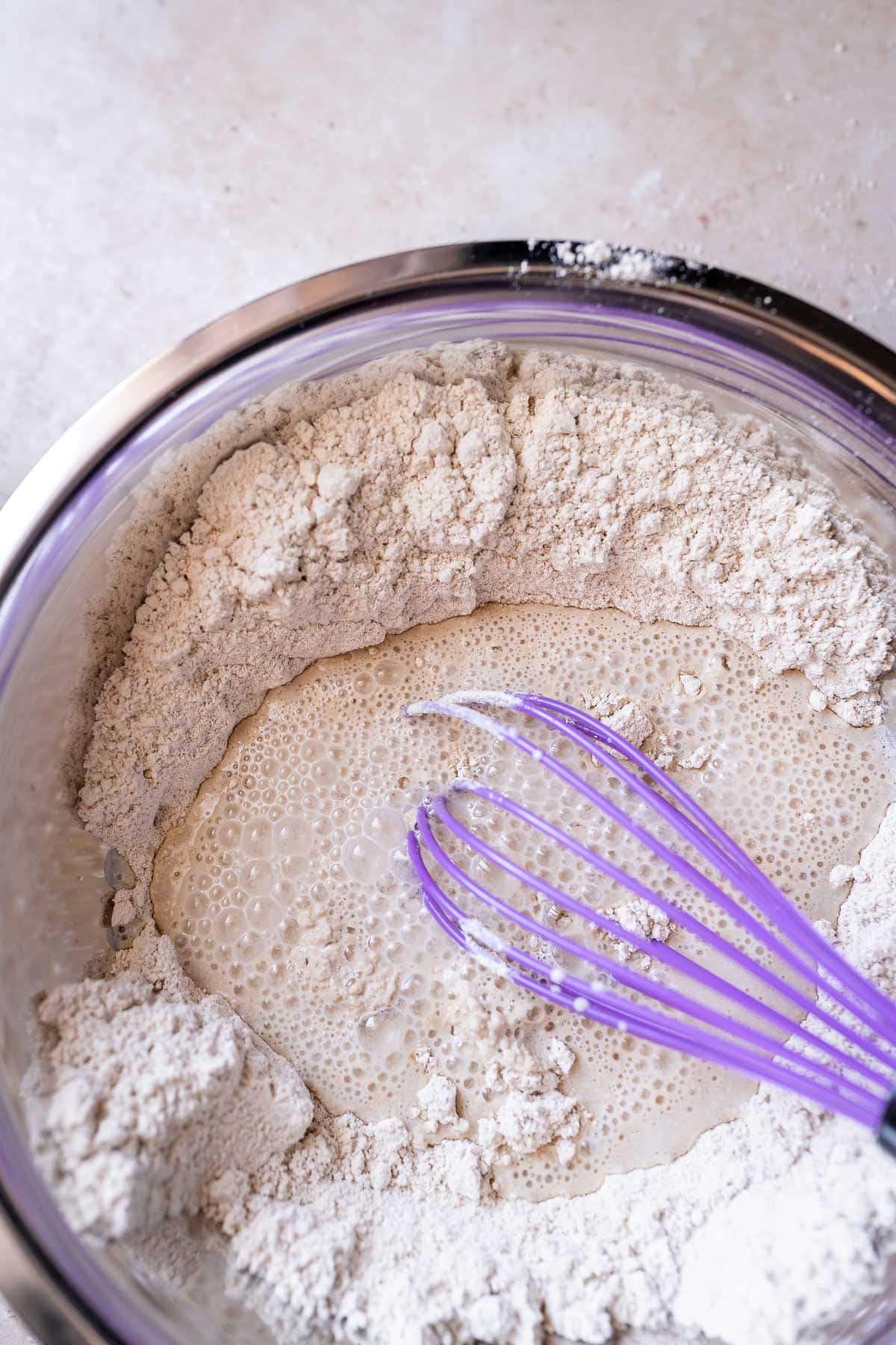 A mixing bowl with dry and wet ingredients and a purple whisk.