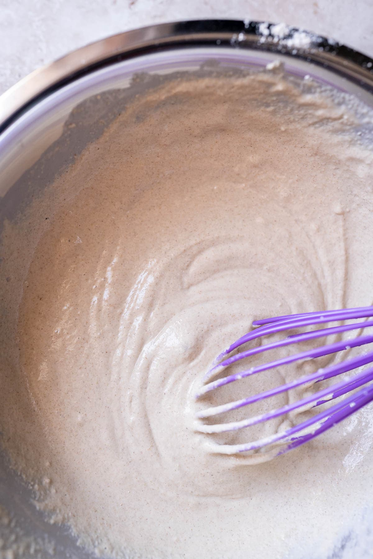 Freshly mixed pancake batter in as silver bowl with a purple whisk.