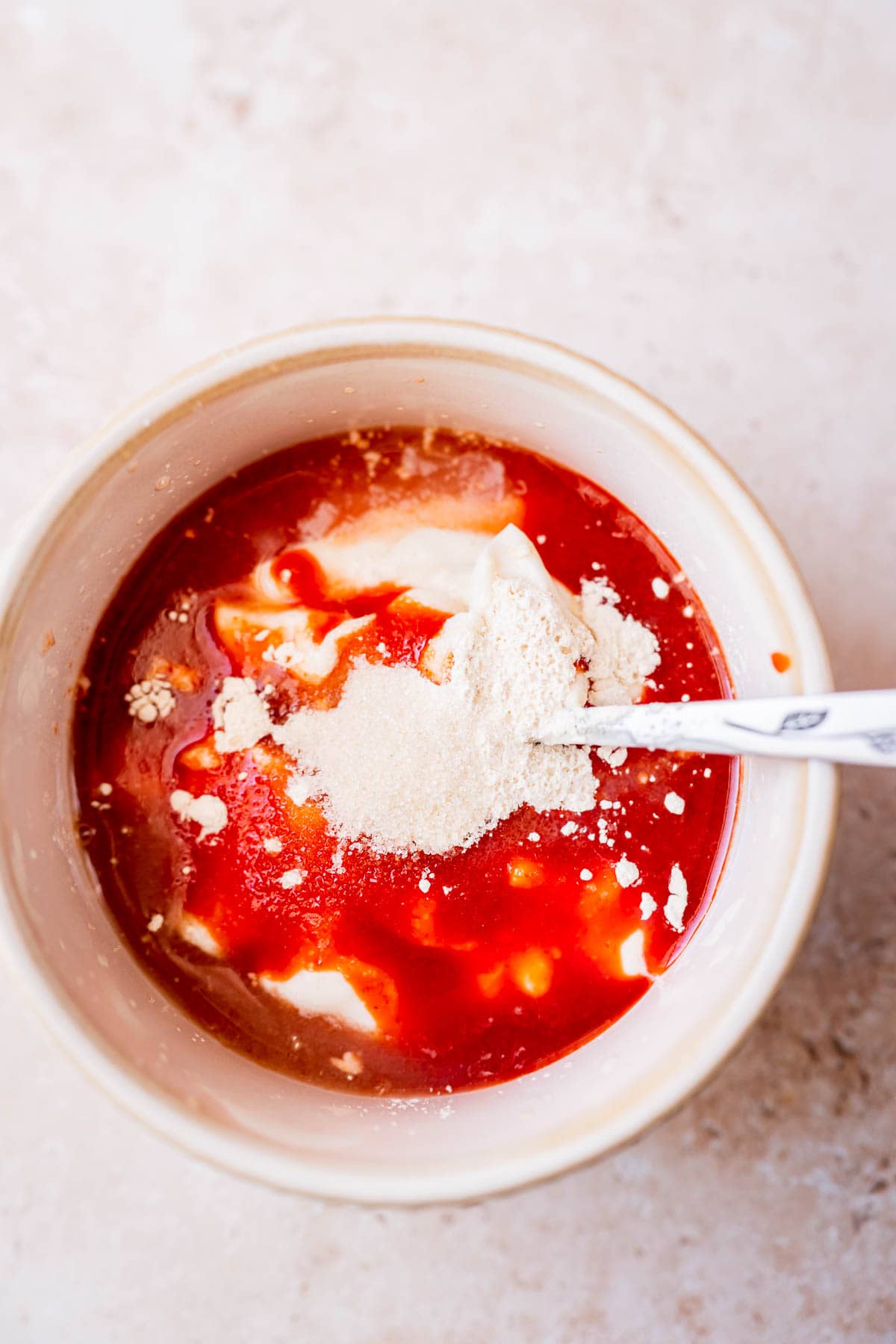 A spoon rest in a small tan ceramic bowl filled with a white sauce, a bright red sauce and a white powder.