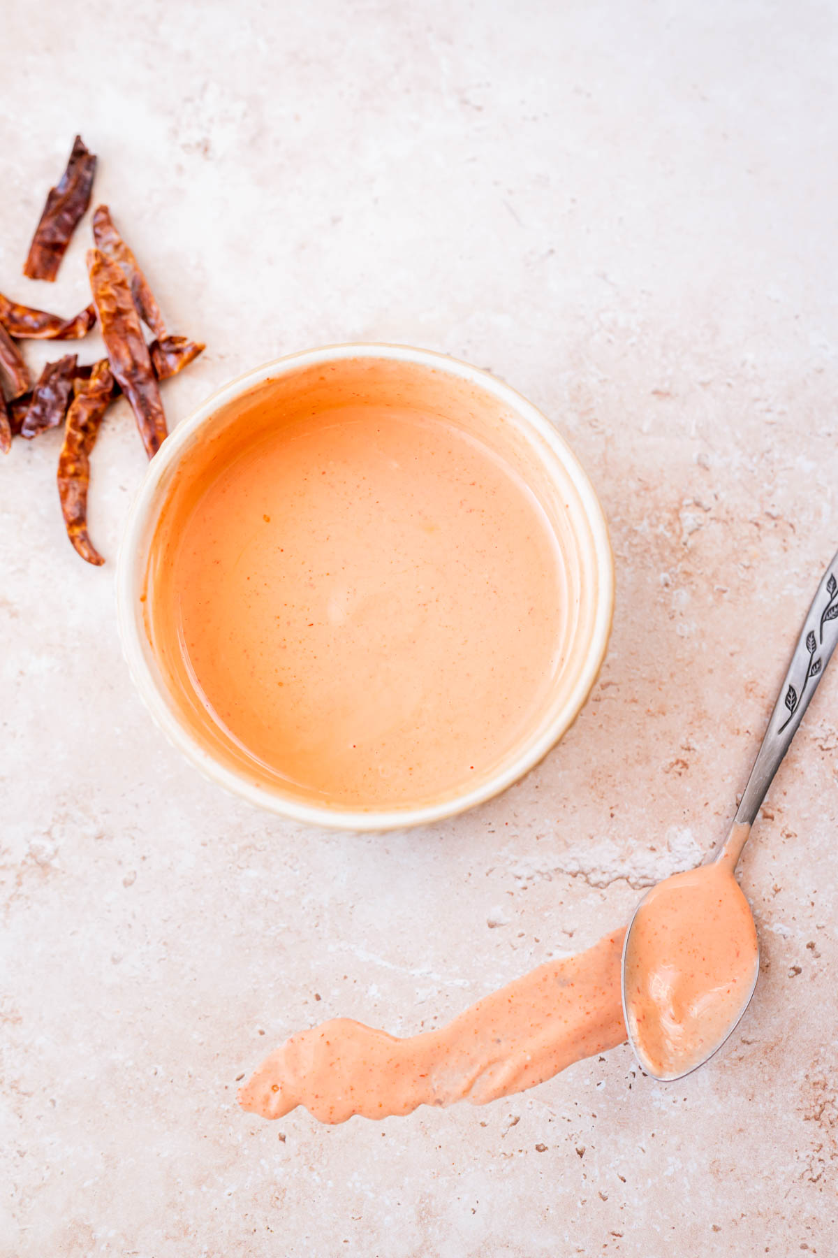 A small tan bowl of spicy aioli rests next to dried peppers and silver spoon.