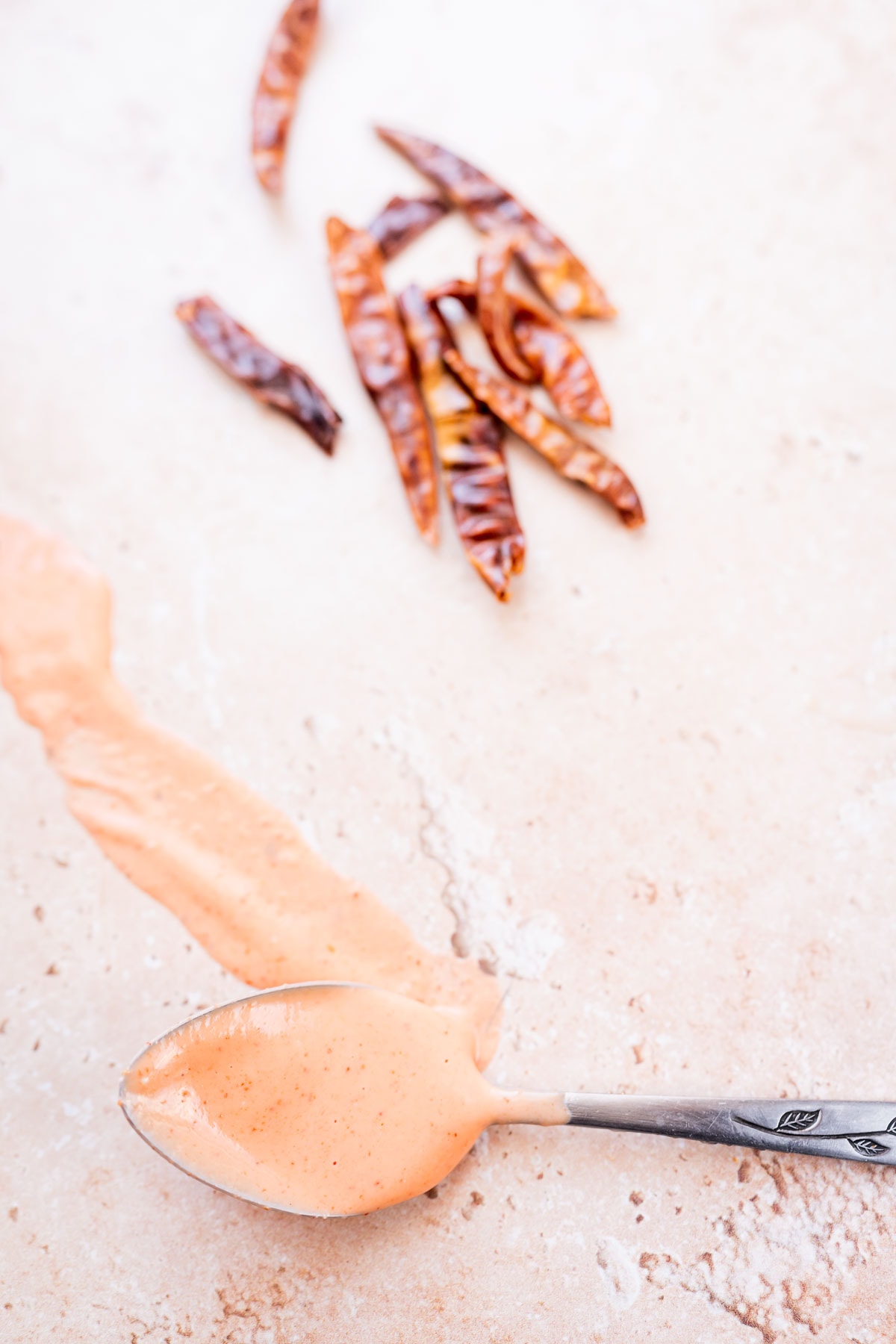 Close shot of a silver spoon drizzling a red sauce on a tan table.