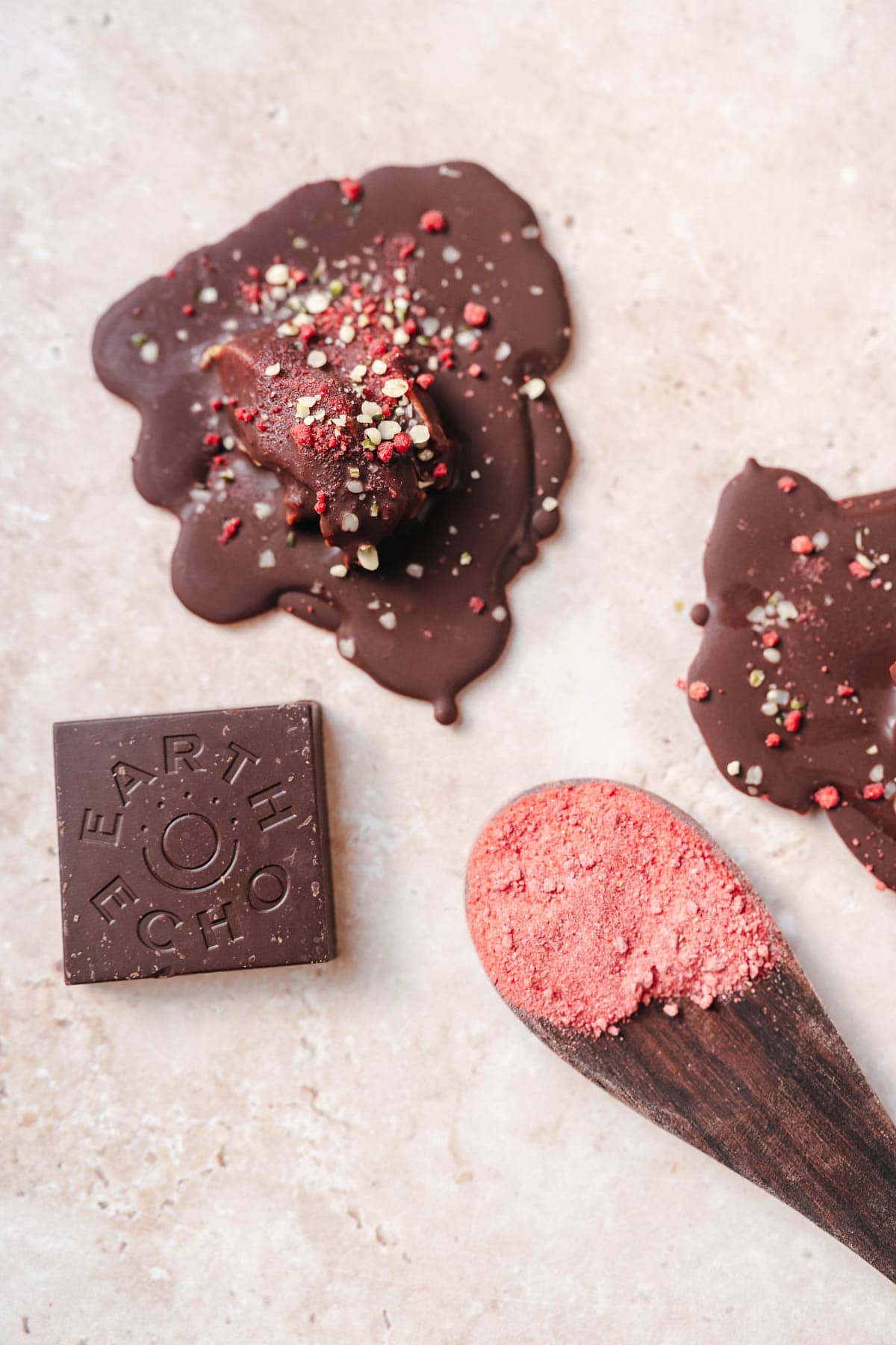 A wooden spoon of strawberry powder rests next to a small chocolate bar that says Earth Echo and homemade chocolate puddles.