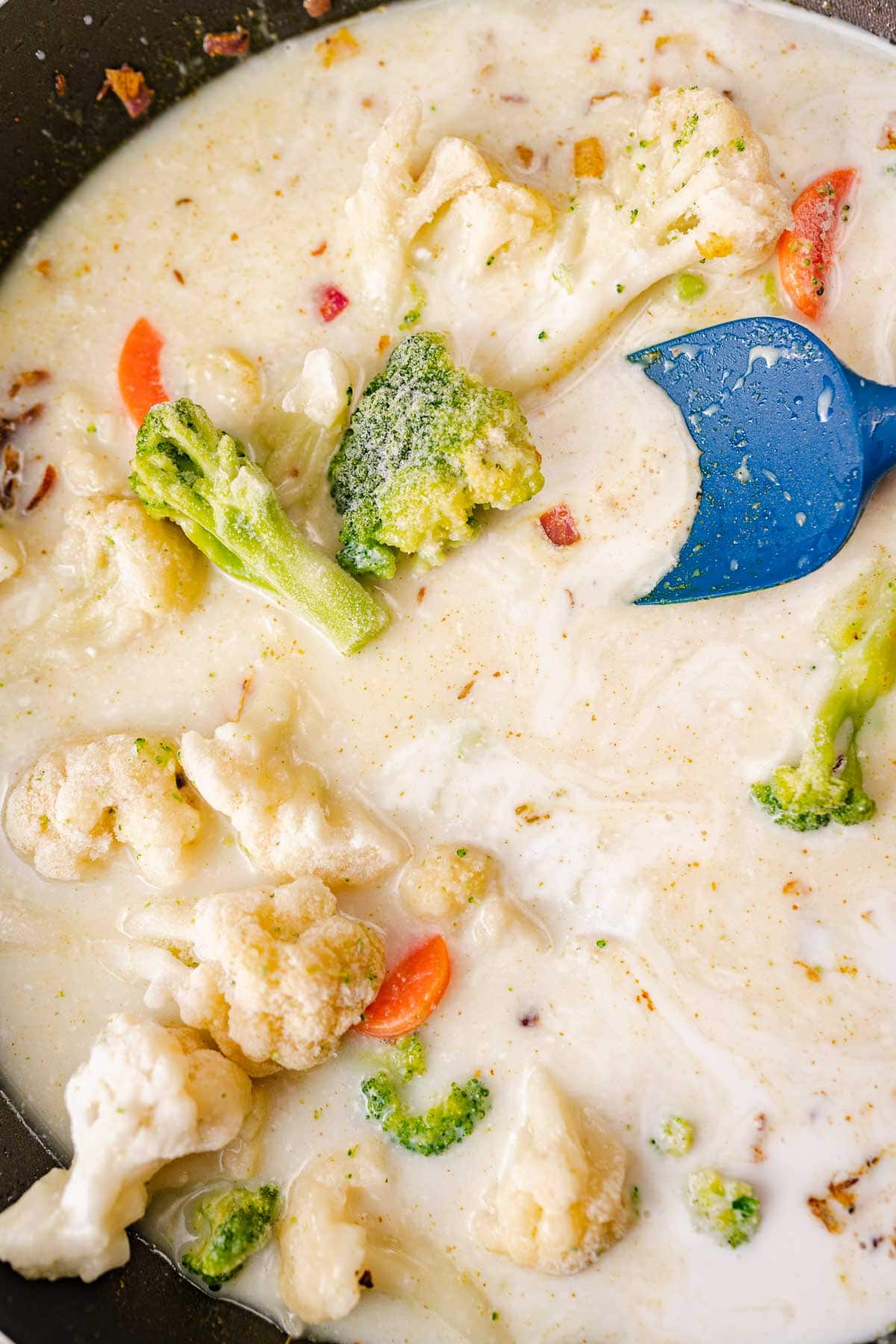 A blue spatula in a skillet with vegetables in a white liquid.