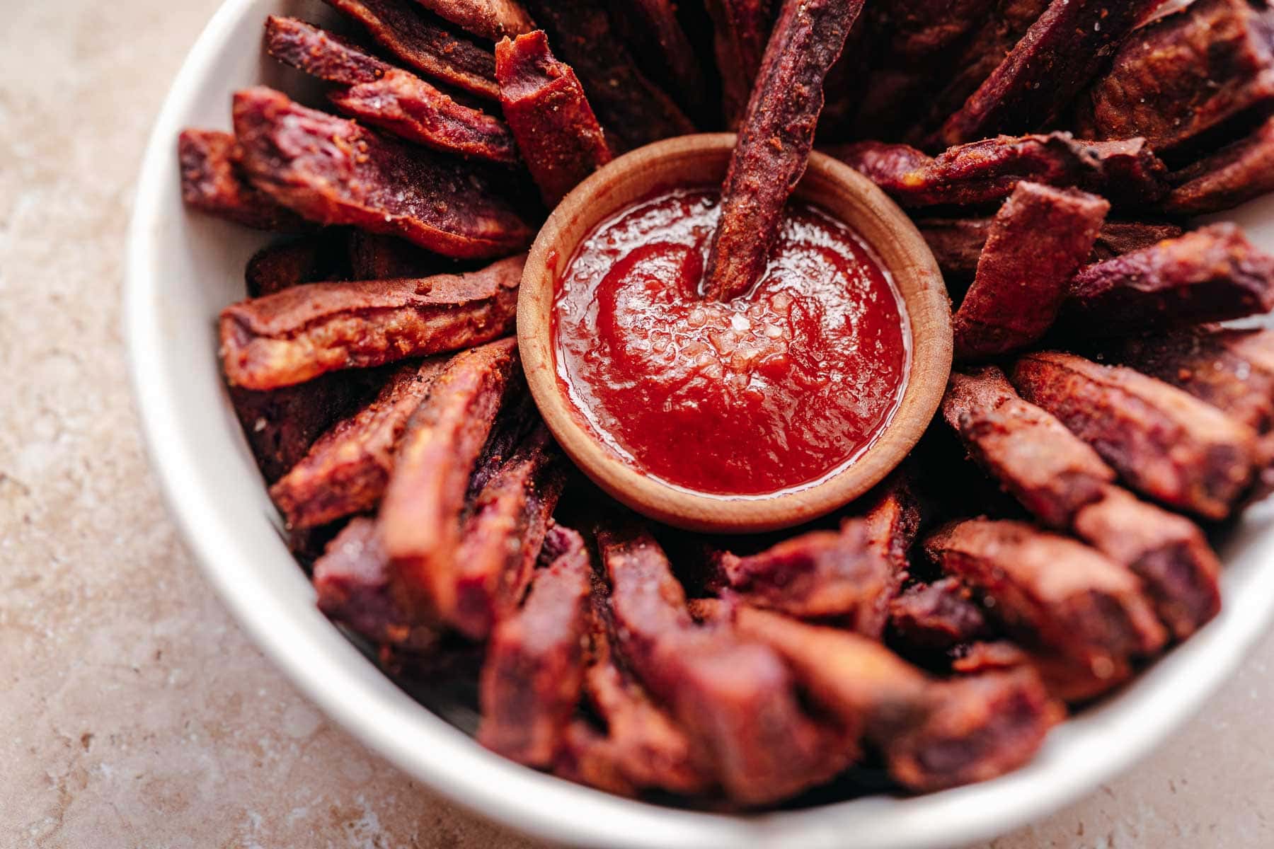 A white ceramic bowl filled with purple sweet potato fries and a small wooden bowl of ketchup.