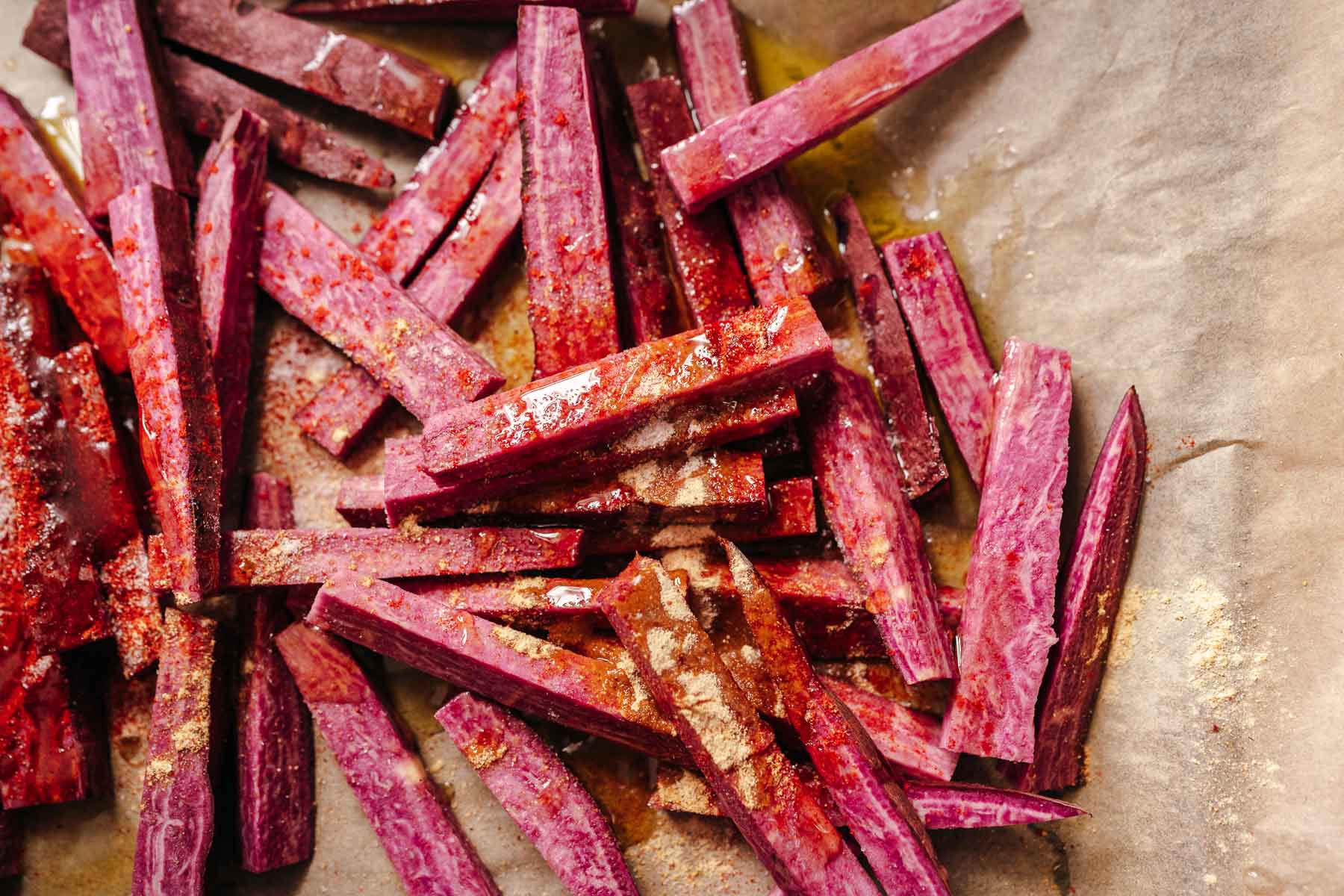 Uncooked purple sweet potatoes drizzled with oil and spices resting on a brown parchment paper.