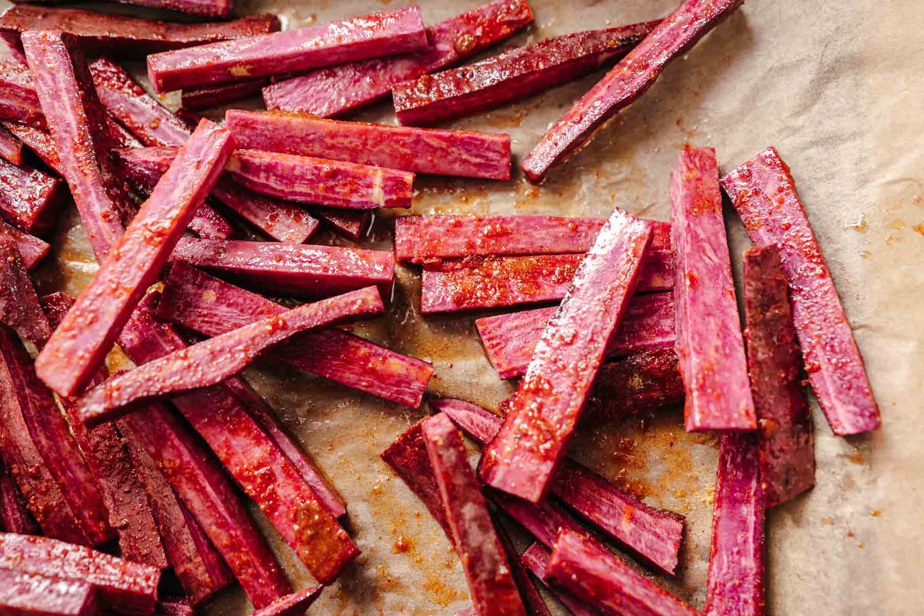 Uncooked purple sweet potato fries tossed with oil on a parchment lined baking sheet.