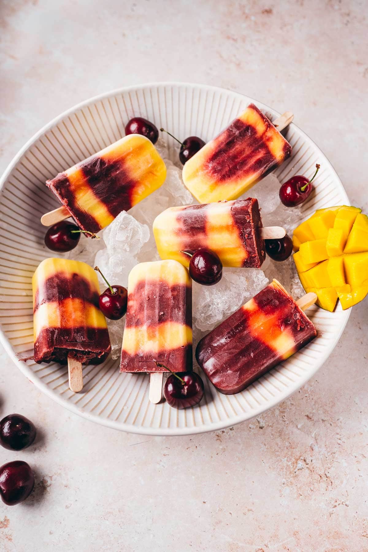 Layered mango cherry popsicles resting a large tan ceramic bowl filled with ice.