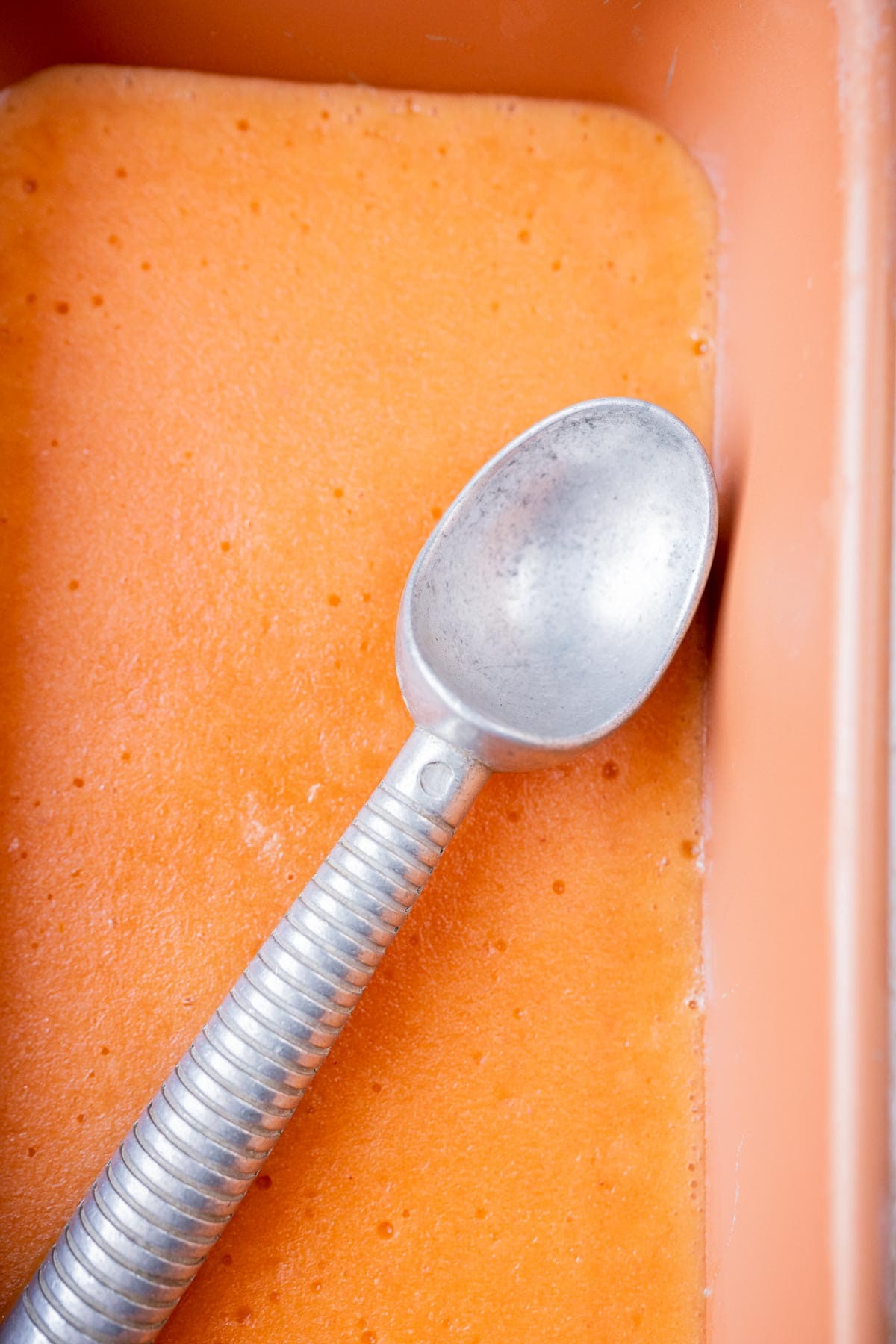 A loaf pan of homemade peach sorbet topped with a silver ice cream scoop.