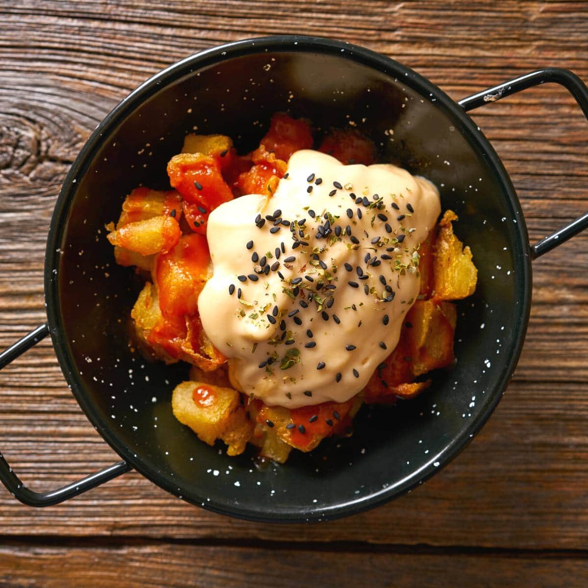 Patatas bravas in a black dish on a wooden table.