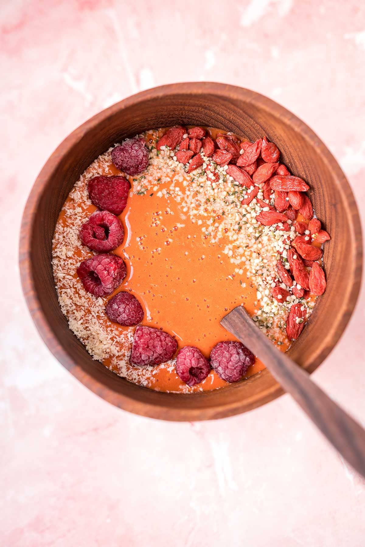Top view of a wooden bowl filled with a dark orange smoothie topped with fruit and seeds.
