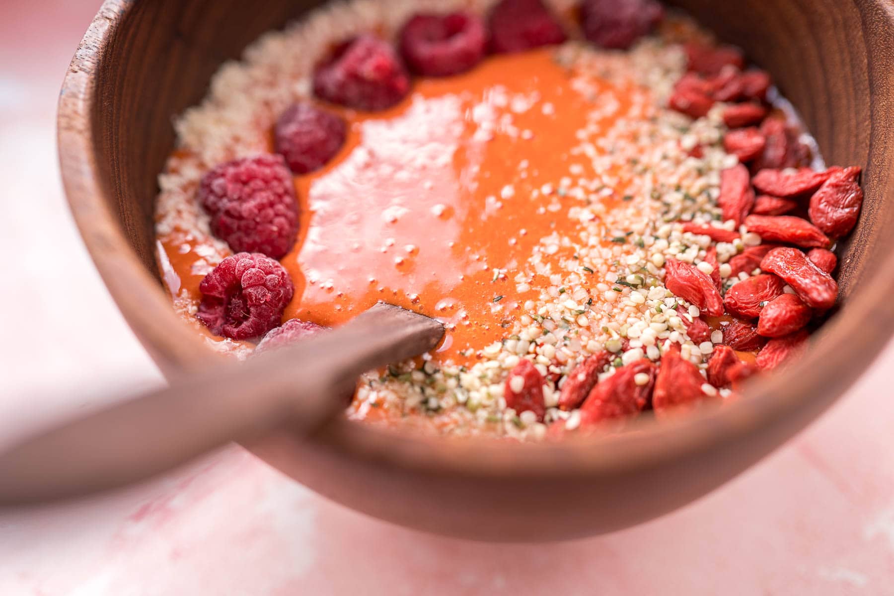 A side shot of a light red goji berry smoothie in a wooden bowl topped with fresh fruit and seeds.