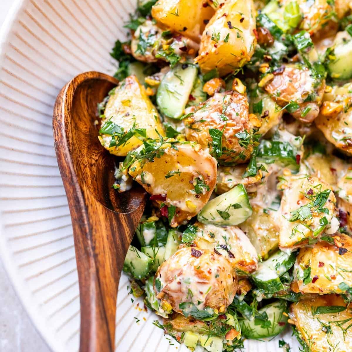 A close shot of a roasted potato salad in a large white bowl with a wooden spoon.