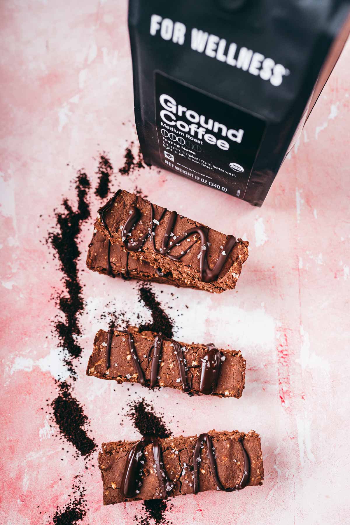 A stack of chocolate protein bars resting on a pink table next to a black bag of ground coffee.