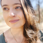 A woman with long hair and a necklace in front of a tree.
