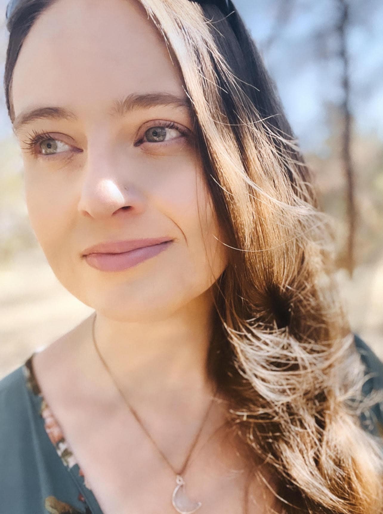 A woman with long hair and a necklace in front of a tree.