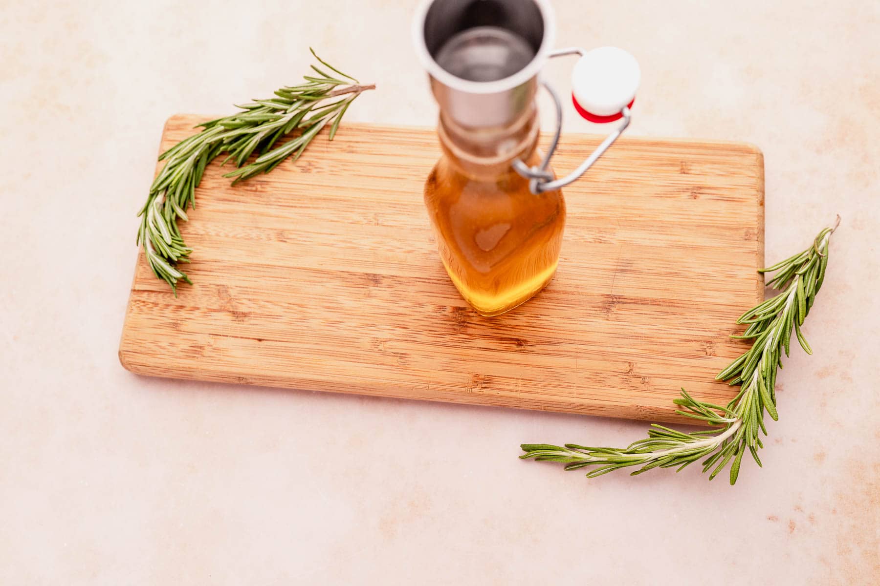 A bottle of olive oil with rosemary sprigs on a wooden cutting board, accompanied by a richly infused rosemary simple syrup.