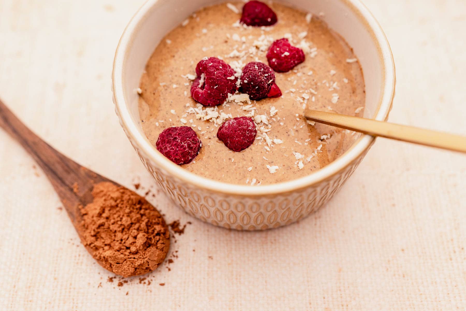A cup of blended chocolate chia pudding topped with raspberries and accompanied by a wooden spoon.