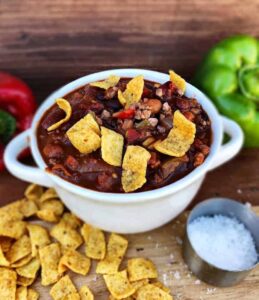 A bowl of chili with tortilla chips on a wooden cutting board.
